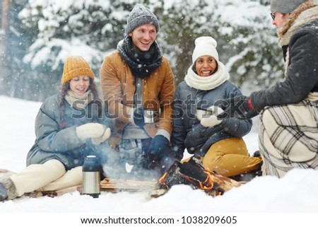 Joyful young people in winterwear sitting in snow arounf fire and having hot drinks