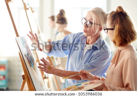 Experienced teacher consulting one of students about her painting during lesson in arts school