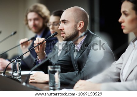 One of young speakers talking in microphone at business or political conference between colleagues