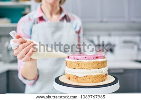 Unrecognizable female pastry cook squeezing strawberry cream on appetizing layer cake in kitchen