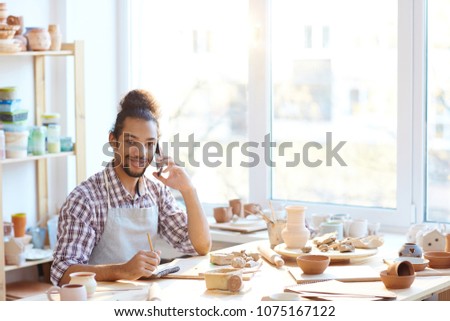 Portrait of young mixed race male potter sitting at workshop table, talking on smartphone and smiling at camera joyfully