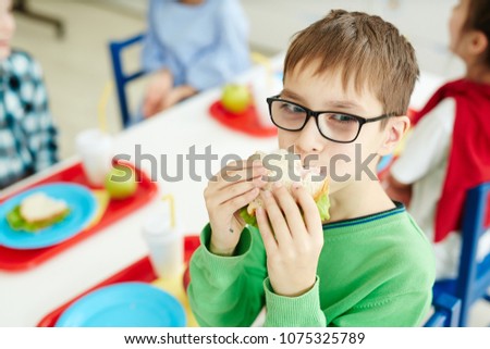 Little Caucasian boy in glasses eating sandwich with appetite while sitting at table with classmates in primary school cafeteria