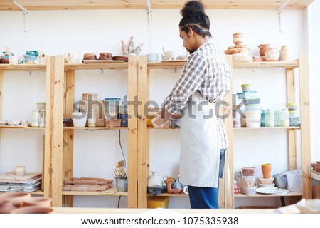 Young African American male potter in apron putting workshop in order while standing by shelf with handicrafts and equipment