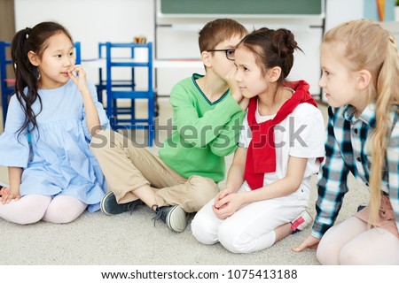 Primary school kids whispering words to each other while playing telephone game in classroom after lessons