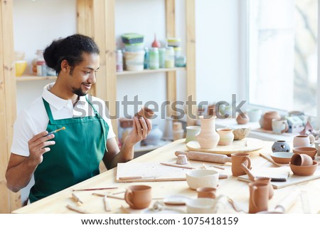 Young mixed race male potter sitting at workshop table, applying glaze on handmade clay pot with paintbrush and smiling