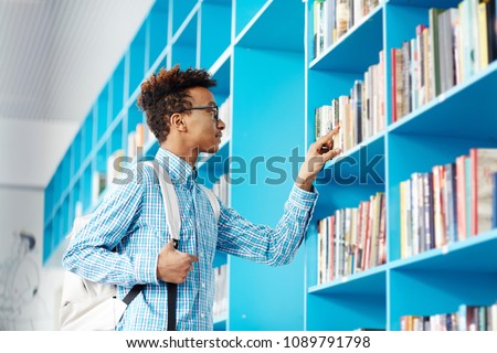 Clever teenage guy with backpack looking for necessary book or manual in college library