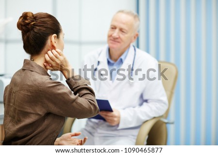 Rear view of nervous female patient with hair bun scratching neck while being interviewed by doctor in clinic