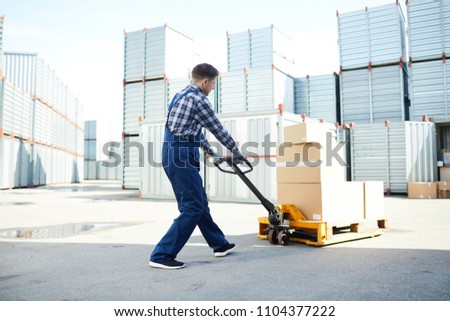 Young worker pushing load cart in front of himself while walking towards storage container