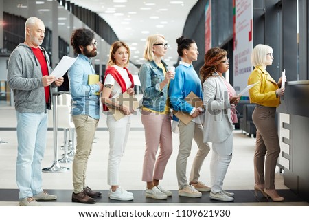 Group of modern multiethnic people of different ages wearing casual clothing holding papers and sketchpads while standing in long line for registration