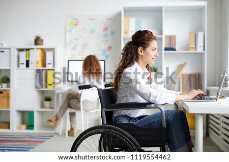 Young disable office worker in wheelchair sitting by desk in front of laptop and networking