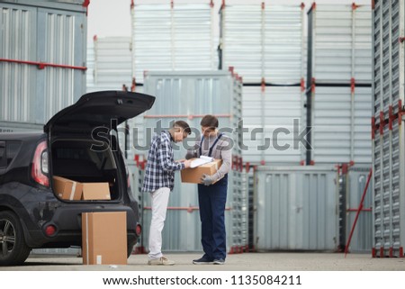 Concentrated young man in casual clothing signing contract with storage company after discussing deal: container storage worker holding box with clipboard and pointing where signature place is