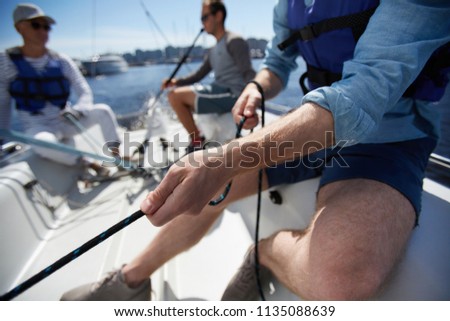 Close-up of male hands pulling rope of sailboat while mooring yacht and sitting with friends on deck