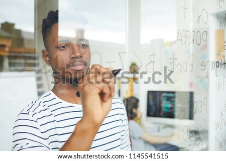 Serious concentrated handsome young African-American web developer using programming language for coding and writing it on glassy wall in office