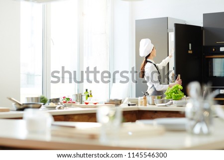 Rear view of busy female chef in hat and apron opening refrigerator and taking ingredients out of it while preparing for cooking in kitchen