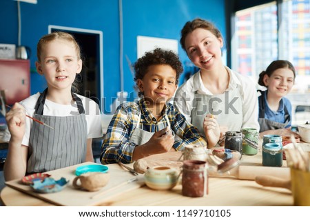 Group of intercultural pupils and their teacher in aprons sitting by wooden table in studio of arts and crafts and painting