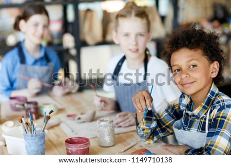 Happy mixed-race boy with paintbrush looking at you at lesson of art and craft