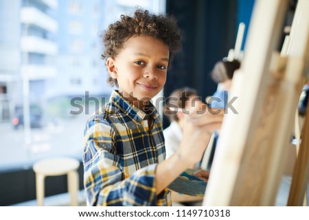 Happy mixed-race pupil looking at camera while standing by easel and painting at lesson in studio of arts