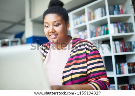 Cheerful confident young African-American overweight student reading online article on laptop while preparing for exam in library