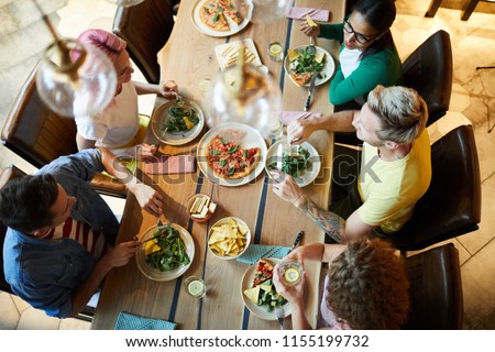 Top view of several young people sitting by wooden table served with tasty food, enjoying dinner and discussing news