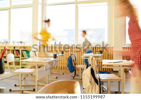 Blurred motion of school children running in classroom, school desks and chairs with hanging satchels in classroom