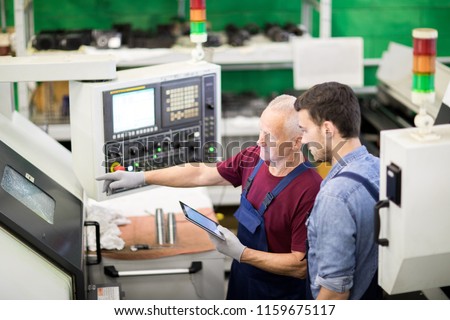 Mature foreman controlling the process of machine on his tablet pc and showing how its work to the worker