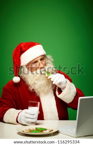 Photo of Santa Claus in eyeglasses looking at laptop while drinking milk with cookies
