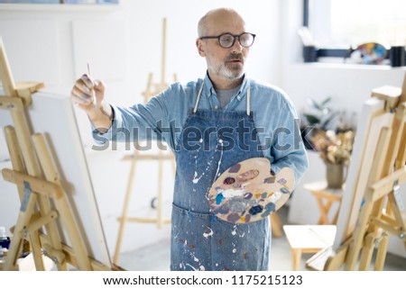 Senior painter in dirty denim apron holding palette while looking at canvas and painting in studio
