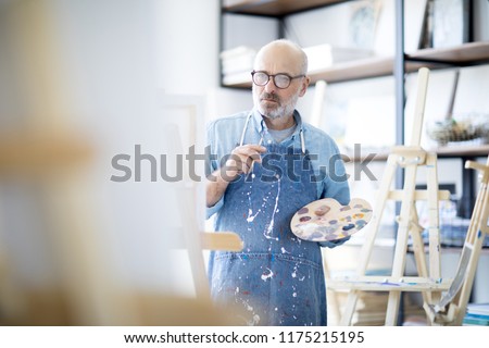 Serious and pensive mature man in apron looking at easel during creative process in studio