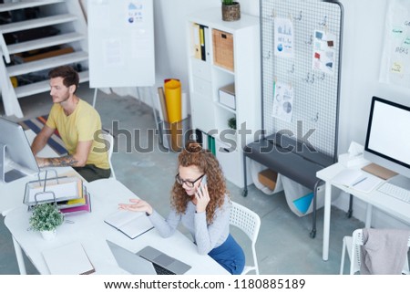 Young secretary or office manager talking by smartphone while sitting by desk in front of laptop