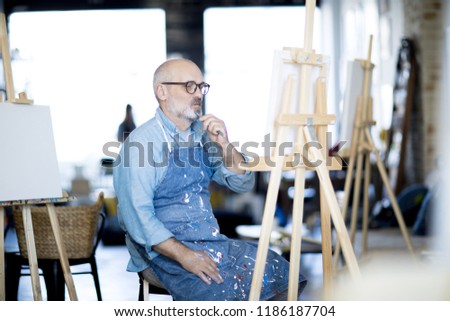 Pensive man in working apron looking at painting and concentrating on suitable color choice