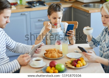 Modern family using mobile gadgets while having breakfast by served table in the kitchen