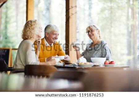 Cheerful optimistic senior friends gathering together in cozy outdoor cafe: they drinking tea with sweets and talking about life