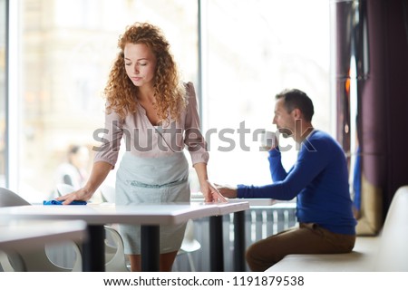 Serious beautiful redhead waitress in apron cleaning in restaurant: she wiping table with cloth while customer drinking coffee behind her