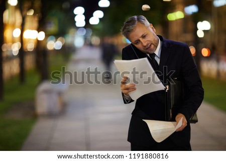 Very busy banker talking by smartphone and looking through financial documents in urban environment after work