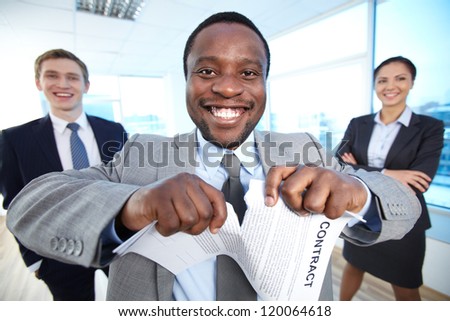Portrait of happy African businessman tearing contract while looking at camera with two partners near by