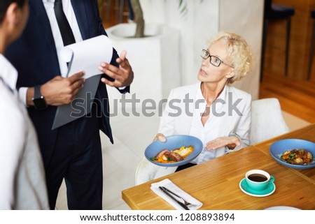 Mature woman sitting at the table during business lunch and complaining on the food to the owner of the restaurant