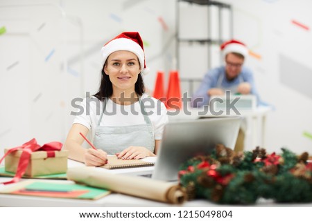 Smiling pretty young lady in Santa hat sitting at table and drawing in sketchpad while creating Christmas design in modern workshop, she looking at camera