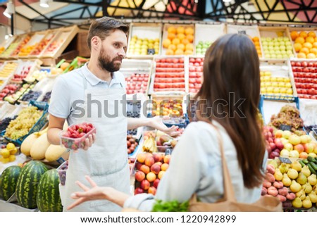 Confused misunderstanding young male grocer in apron holding container of strawberries shrugging shoulders while talking to customer in farmers market