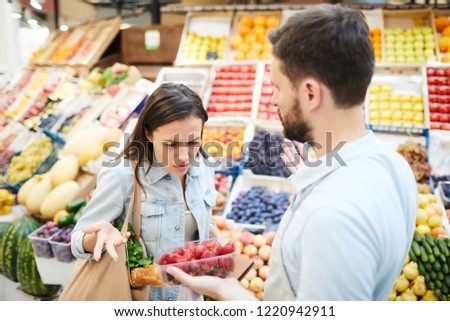 Puzzled frowning young female customer with shopping bag on shoulder displeased with quality of products and looking at strawberry shown by grocer in farmers market