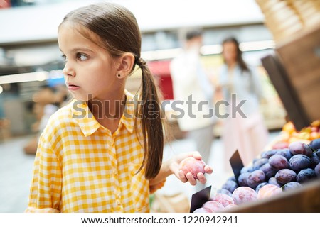 Worried girl with pony tails wearing checkered shirt standing at food shelves and holding plum while stealing it in food store