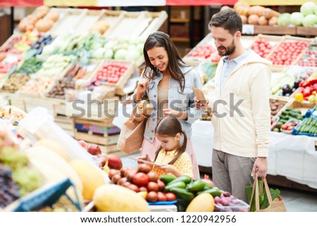 Serious concentrated little daughter with pony tails standing at food shelves and scanning price tag using tablet in food store, cheerful parents looking at tablet screen while making purchases