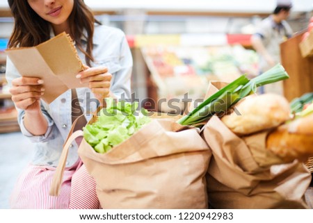 Close-up of content young woman crouching near full shopping bags and reading shopping list in organic food store