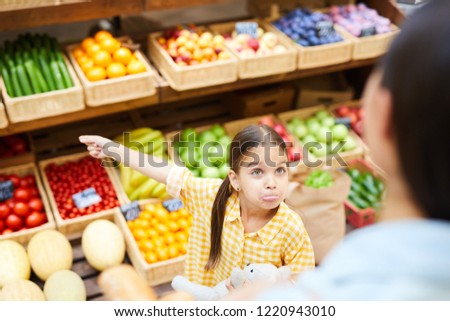 Upset grimacing daughter with pony tails complaining to mom and gesturing aside, she getting hysterical in organic food shop