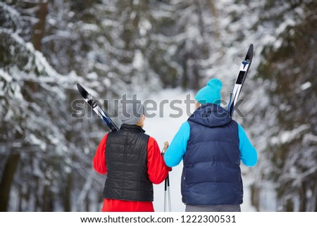 Back view of mature sporty couple in activewear carrying skiing equipment while moving in winter woods
