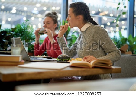Two teenage students sitting in college cafe, eating fast food and watching webcast in laptop