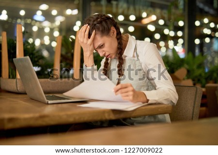 Tired waitress in apron reading one of documents while sitting by table in front of laptop
