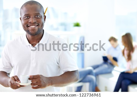 Portrait of happy African guy with cup looking at camera in working environment