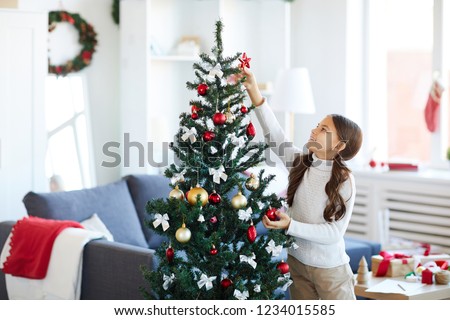Happy girl trying to put red decorative star on top of Christmas tree while decorating it
