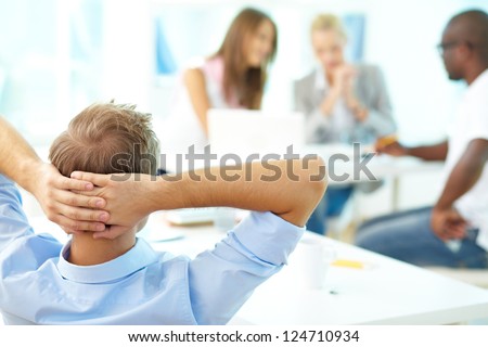 Rear view of relaxed teacher keeping hands behind head and looking at group of students working