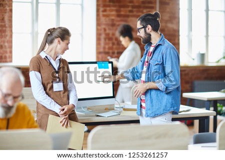 Young confident designer or analyst pointing at computer screen while explaining online statistics to one of colleagues